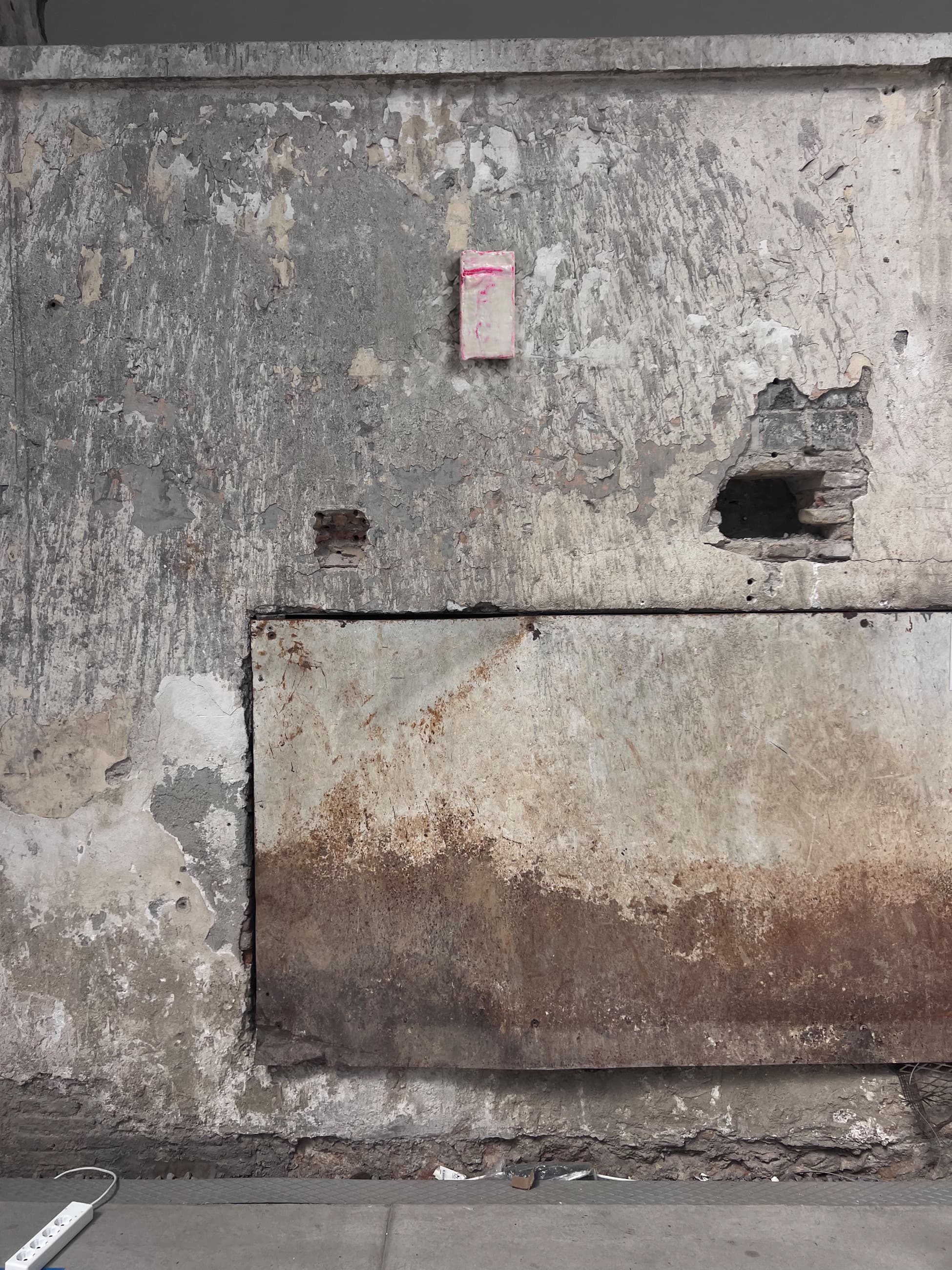 A single brick on the bare concrete wall of the factory, paint and rust around it