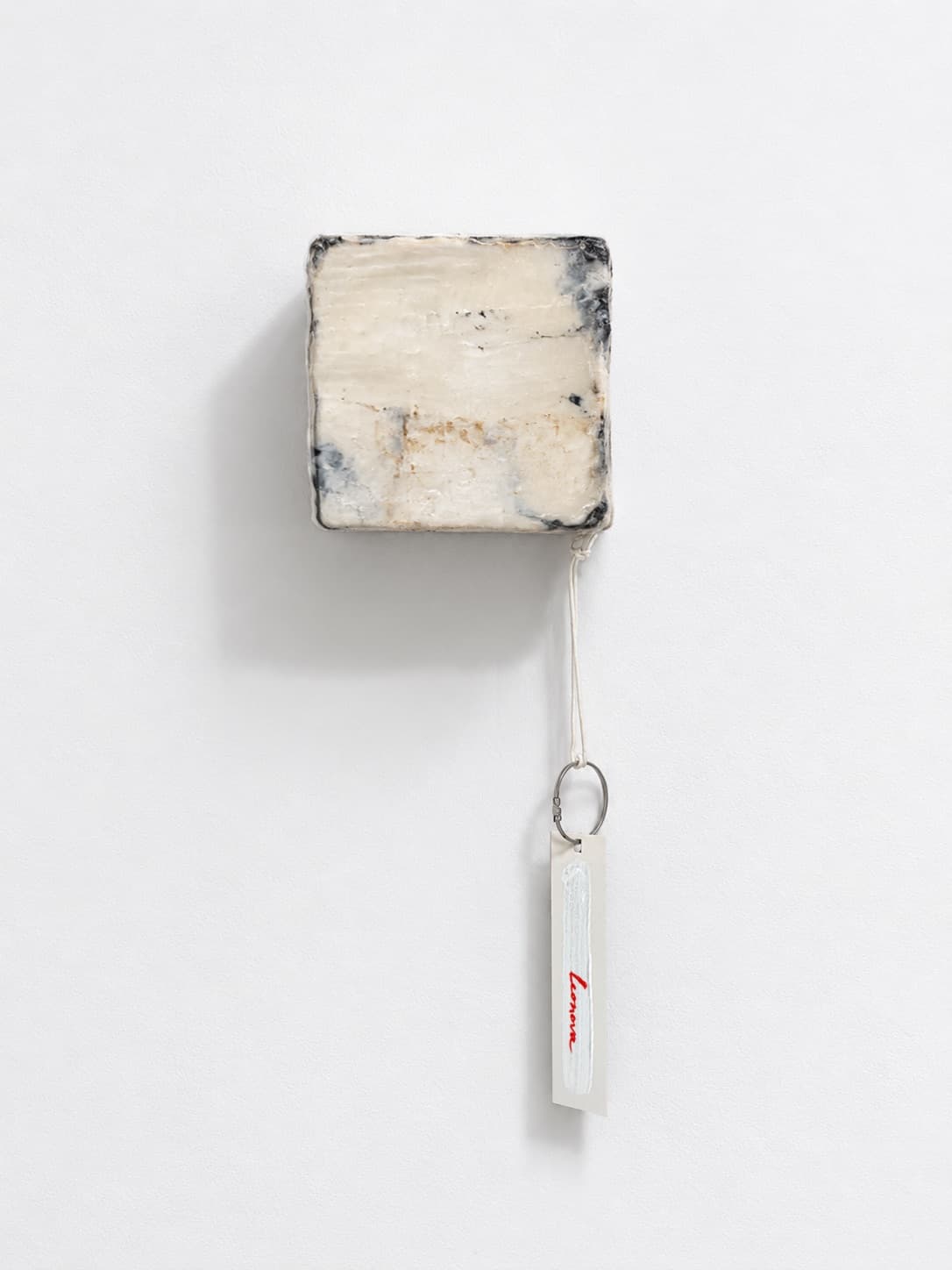 A pale cube of textile waste hung on a white wall, a signed paper tag hanging beneath it on a ribbon