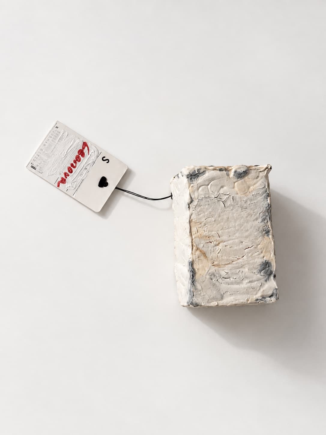 A small white object lying on a pale ground, a signed paper tag on a thread beside it