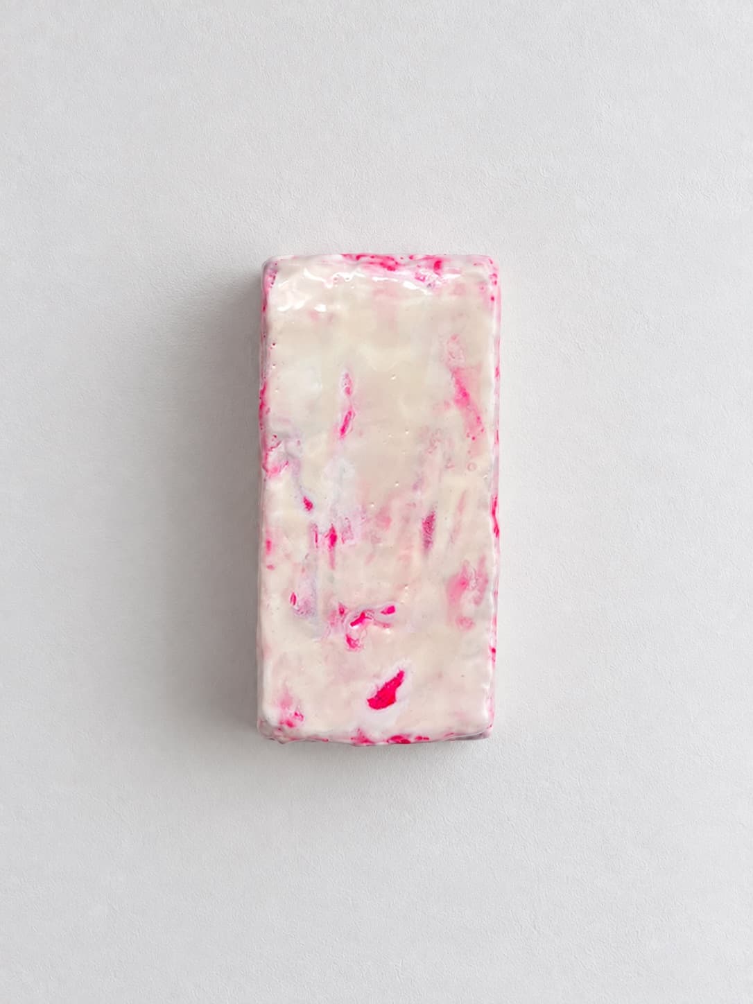A brick of compressed textile waste sealed in white wax with pink showing through, mounted upright on a white wall